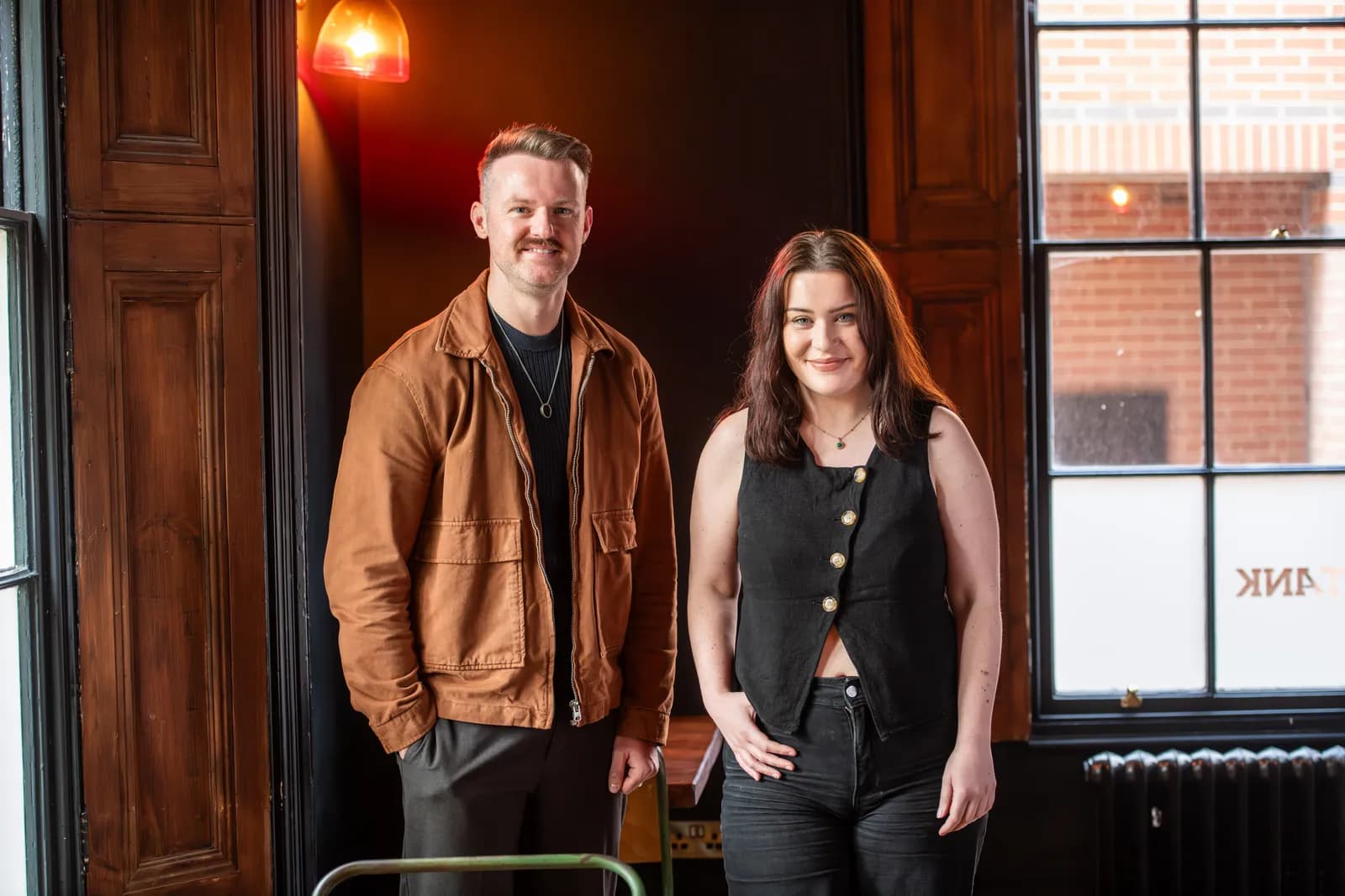 A man and a woman stand next to each other, both smiling at the camera, inside a room with dark wood paneling and a large window.