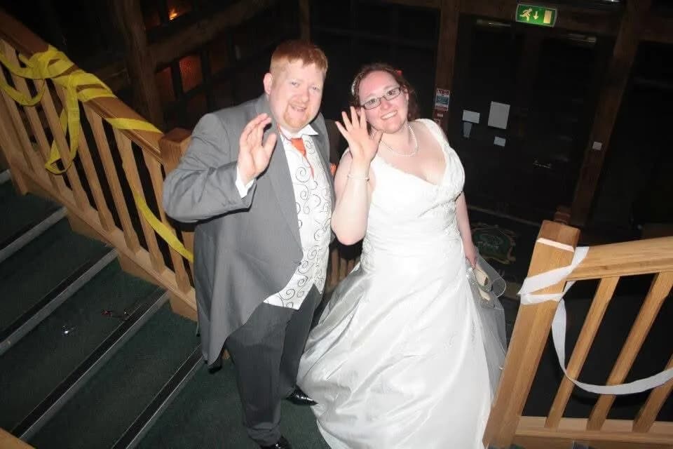A man in a gray suit and a woman in a white wedding gown wave at the camera while standing on a staircase.