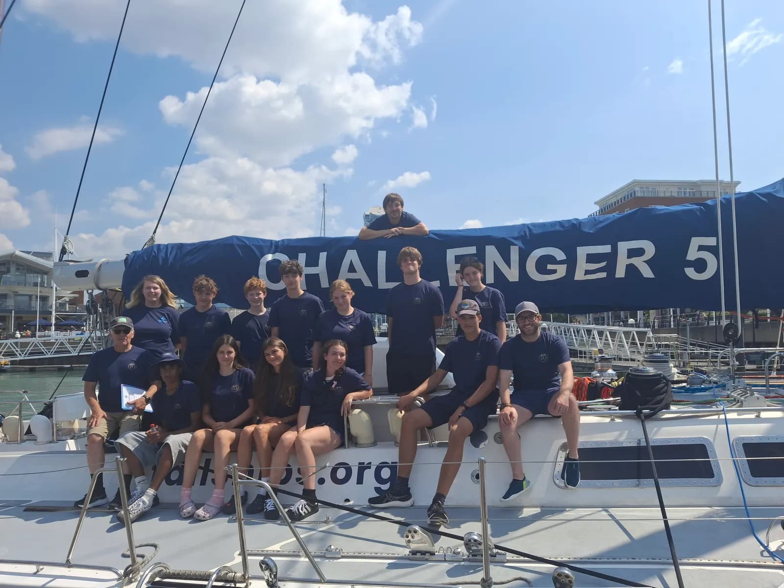A group of young adults and two older men pose for a photo on a sailboat named "CHALLENGER 5" under a blue sky with some clouds.