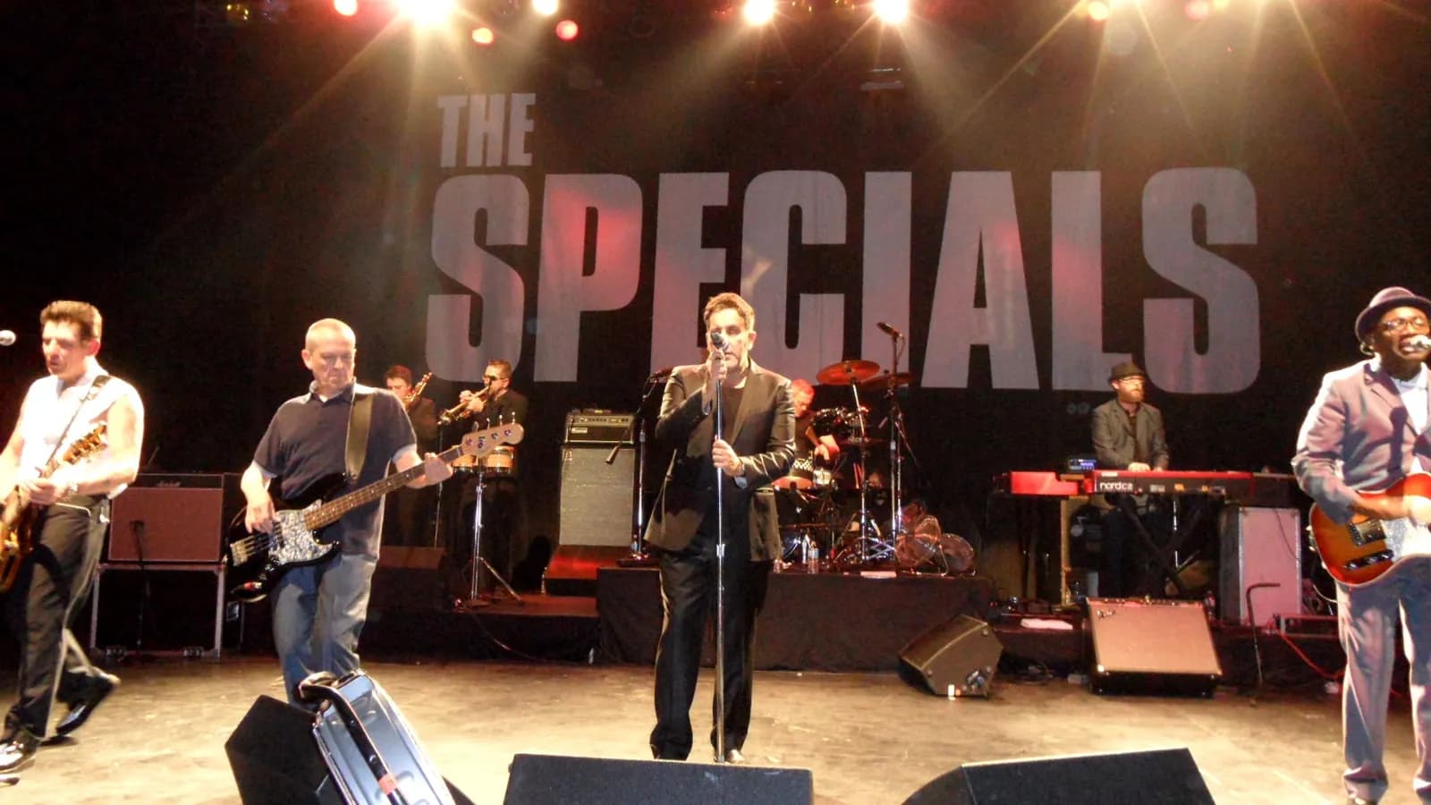Ska band The Specials performing live on stage with a large white band name logo behind them and bright stage lights.