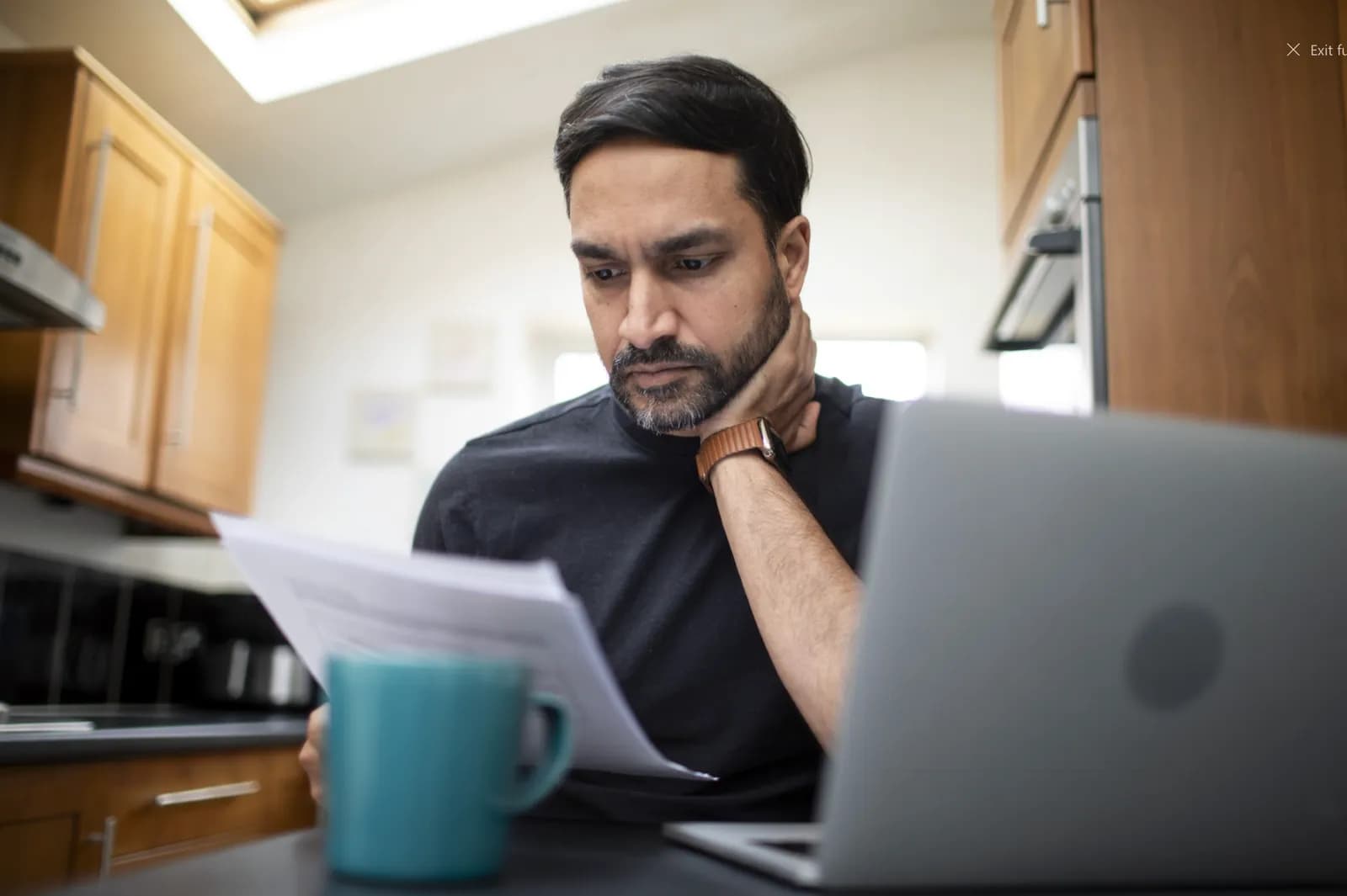 A man wearing a black shirt and a watch on his left wrist holds papers in his hands and looks down at them with a concerned expression, while a laptop and a blue mug sit on the table in front of him.