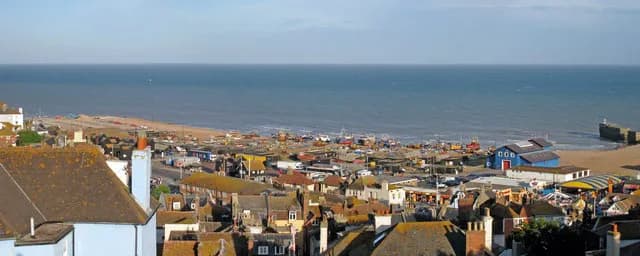 A coastal town with many brown-roofed buildings in the foreground, a sandy beach with boats and blue structures, and the expansive blue sea under a clear sky.