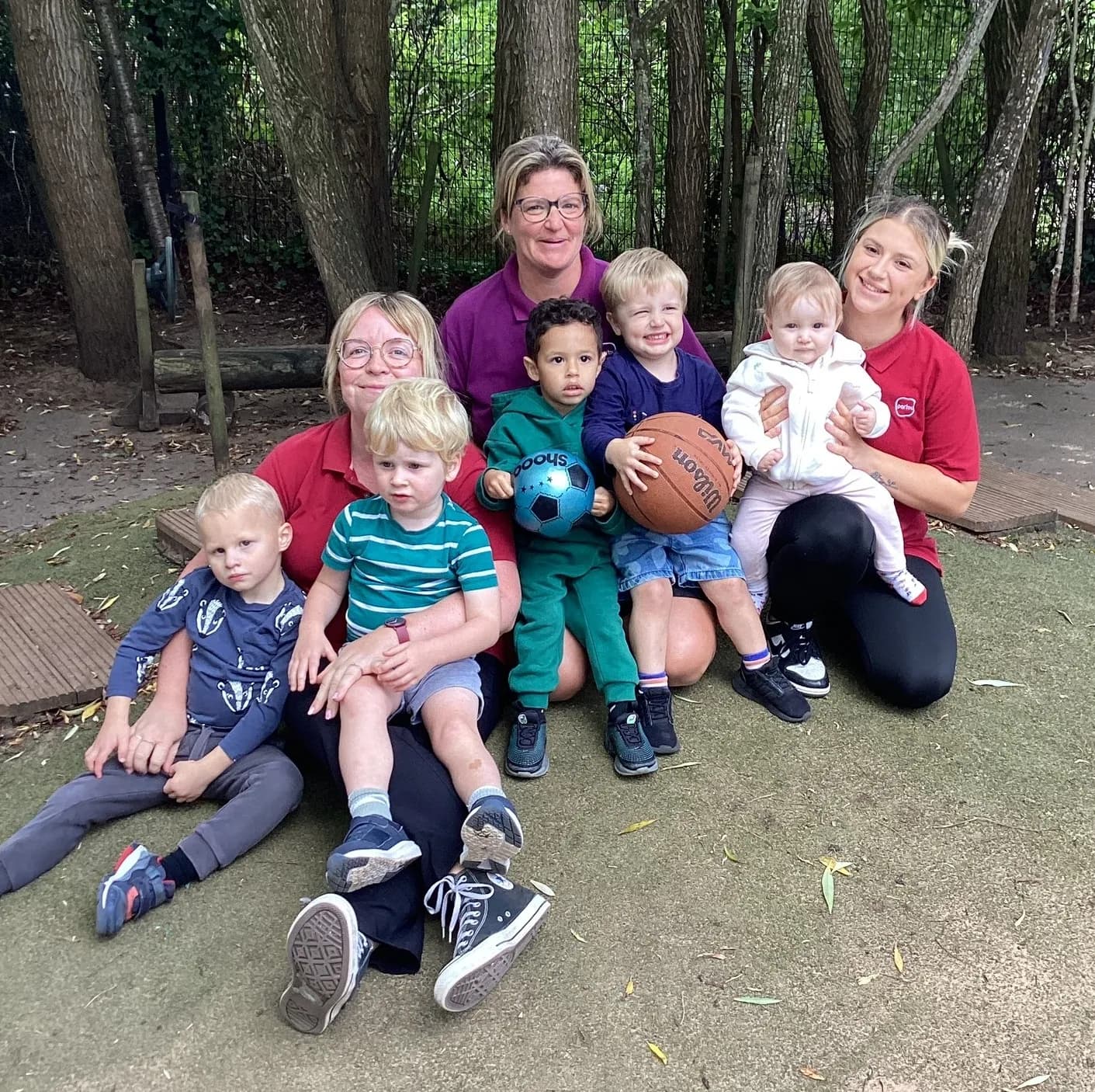 A group of three adult women and six children of various ages are sitting together outdoors, smiling at the camera, with a backdrop of trees and greenery.