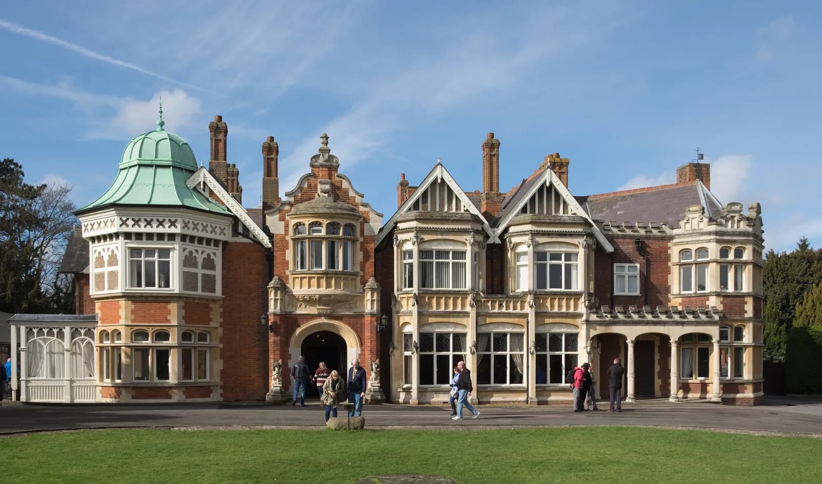 A historic Bletchley Park manor house with intricate brickwork, multiple bay windows, and a distinctive copper dome stands under a bright sky, with a few people walking on the paved area in front.