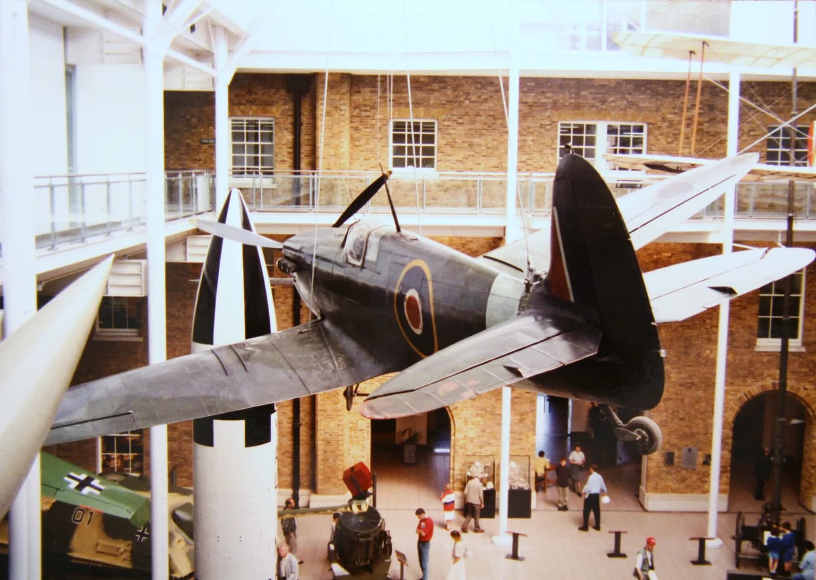 A dark-colored fighter plane with circular markings on its side hangs suspended in a museum hall with brick walls, surrounded by other exhibits and a few visitors.