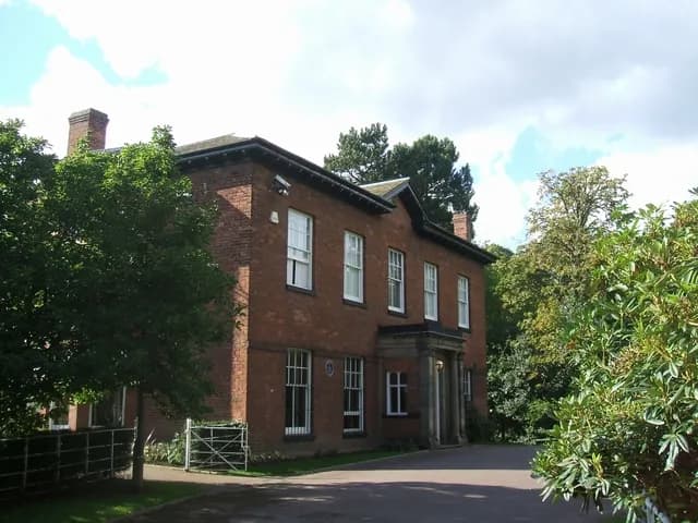 A large red brick house with white-framed windows and a dark portico stands surrounded by green trees under a partly cloudy sky.
