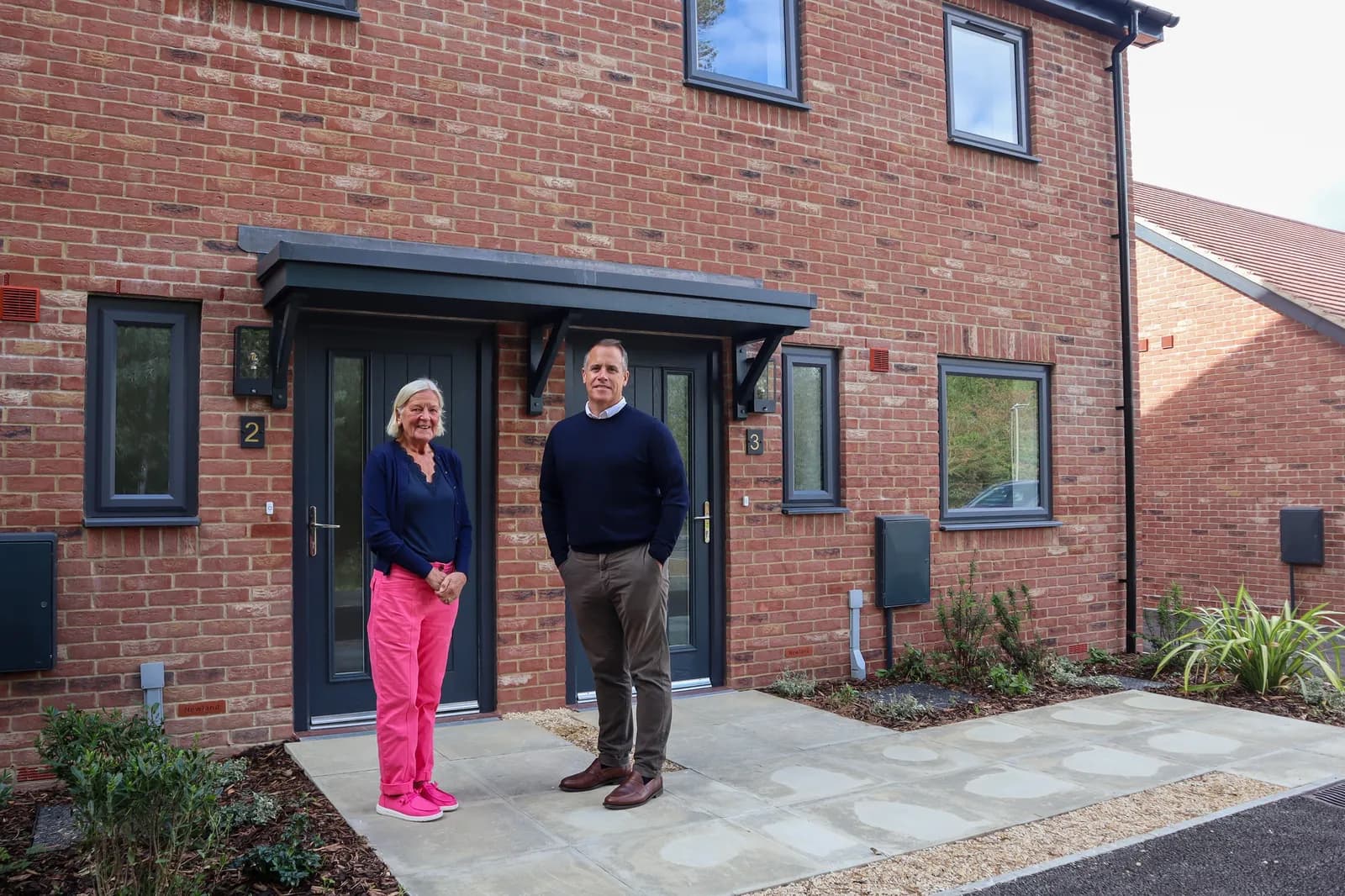 A smiling woman in pink pants and a man in a navy sweater stand in front of two brick townhouses.