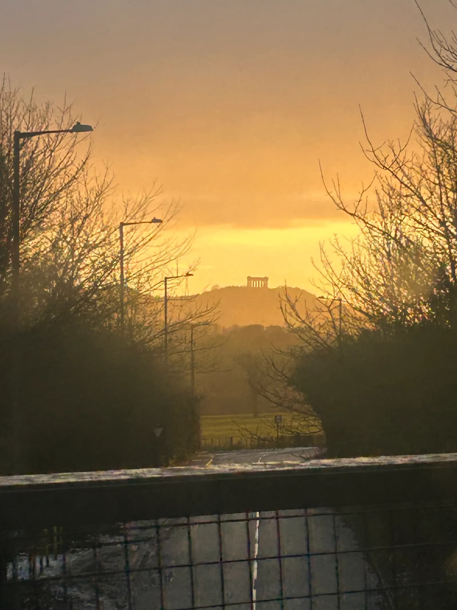 A classical building, likely a monument, is silhouetted on a distant hill under a bright orange and yellow sunset sky, framed by dark, bare trees and streetlights in the foreground.