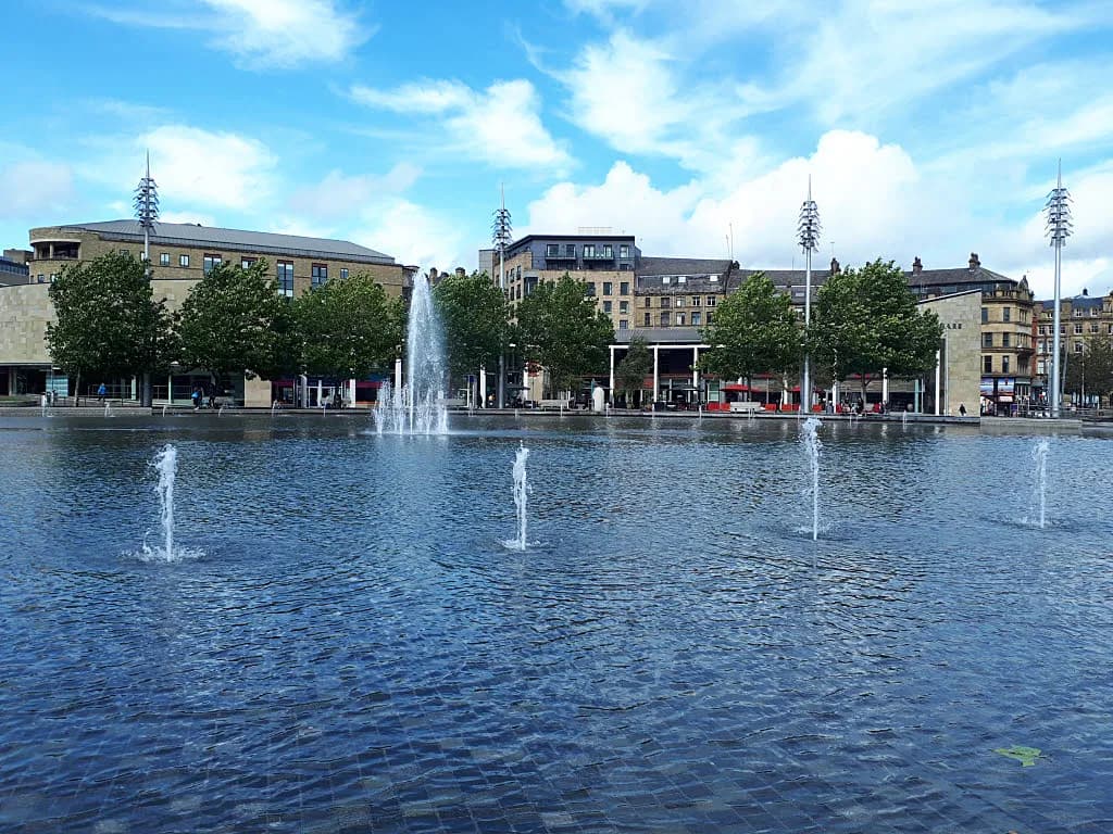 A wide shot of a public square featuring several fountains spraying water into a rectangular pool, with modern buildings and green trees in the background under a blue sky with white clouds.