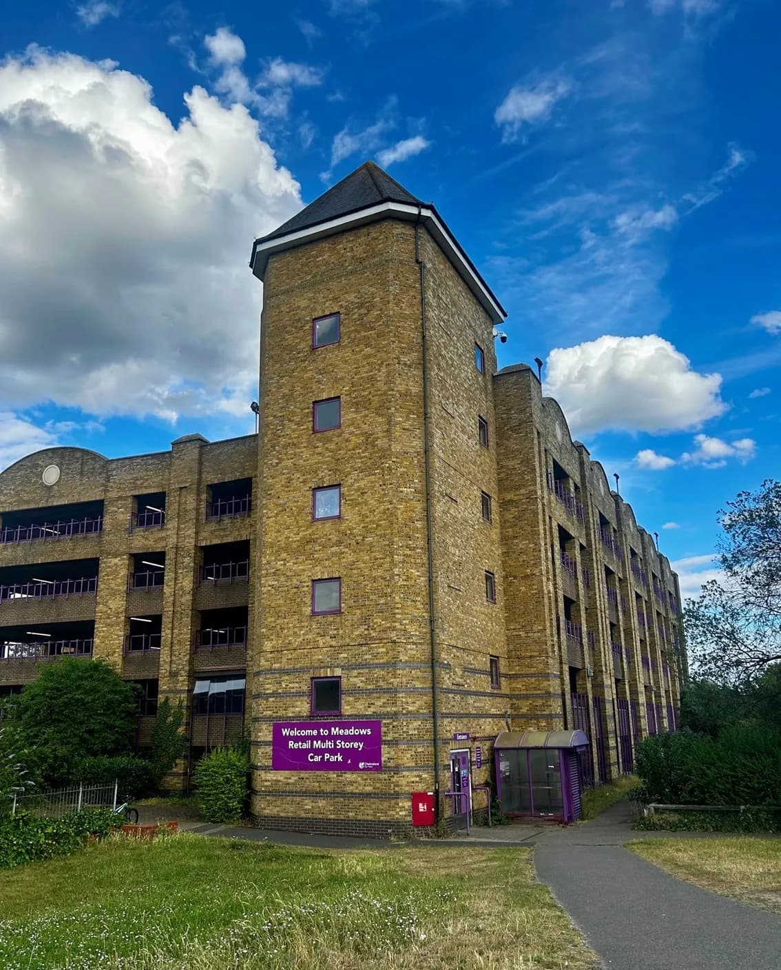 A large brick multi-story car park with a tower-like structure on the left side, set against a bright blue sky with fluffy white clouds.