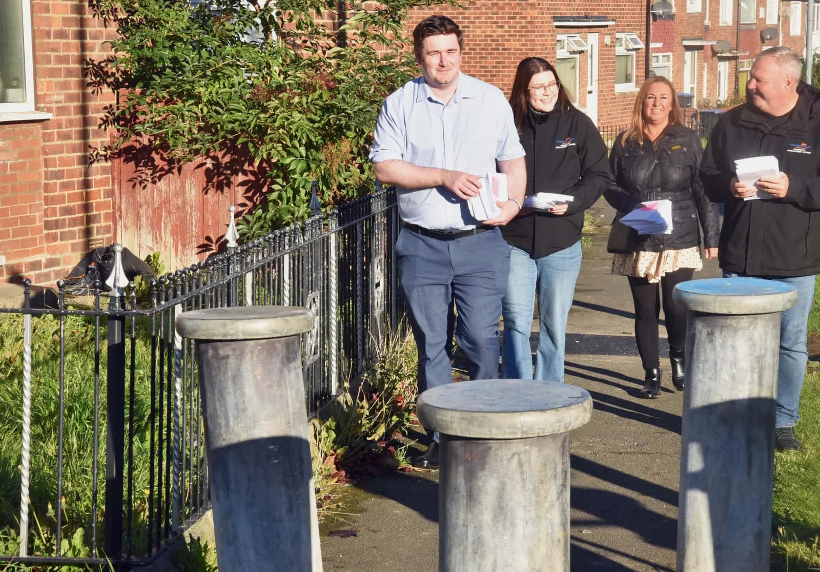 Four people, two men and two women, walk along a sidewalk next to a brick building and a black metal fence while carrying papers.