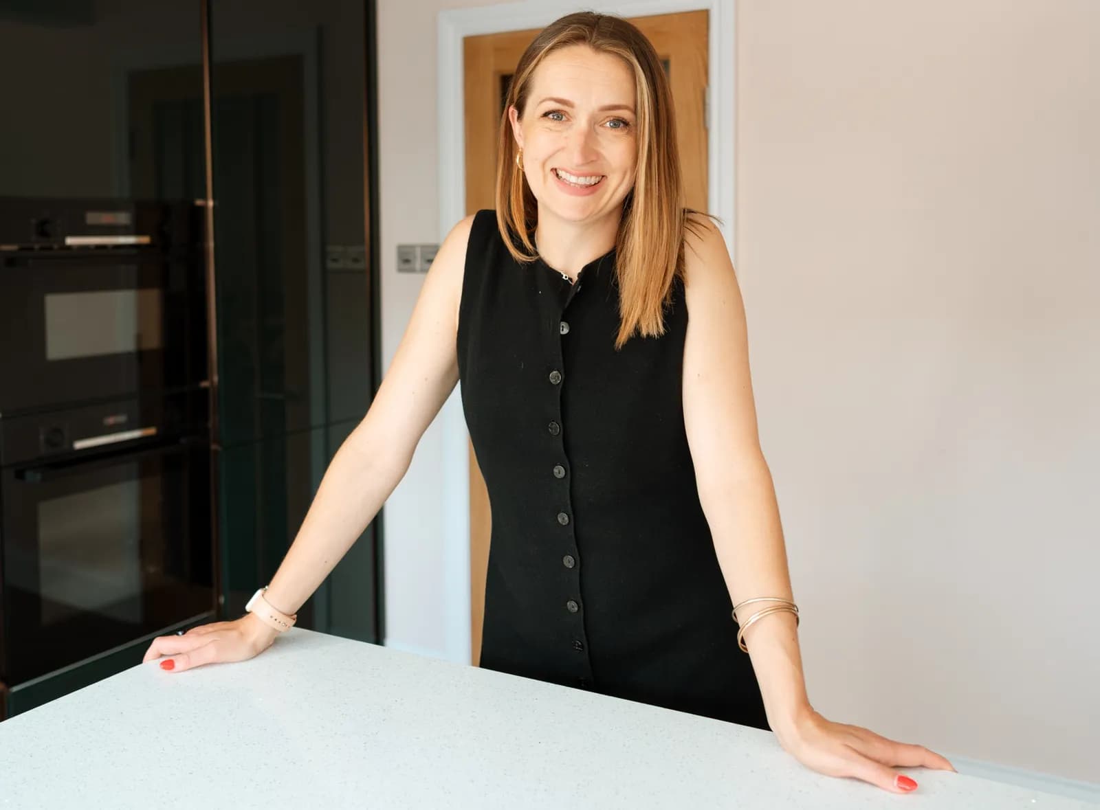 A smiling woman with long blonde hair, wearing a black sleeveless dress, leans on a white countertop in a modern kitchen.