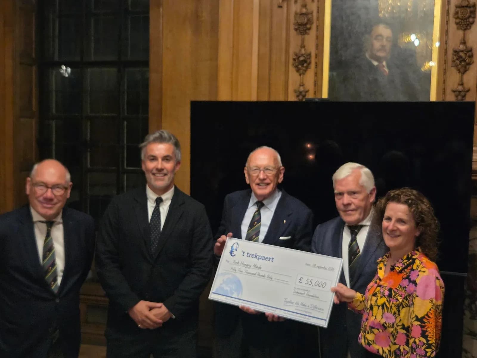 Five adults, three men and two women, stand smiling in a wood-paneled room, holding a large check for £55,000.