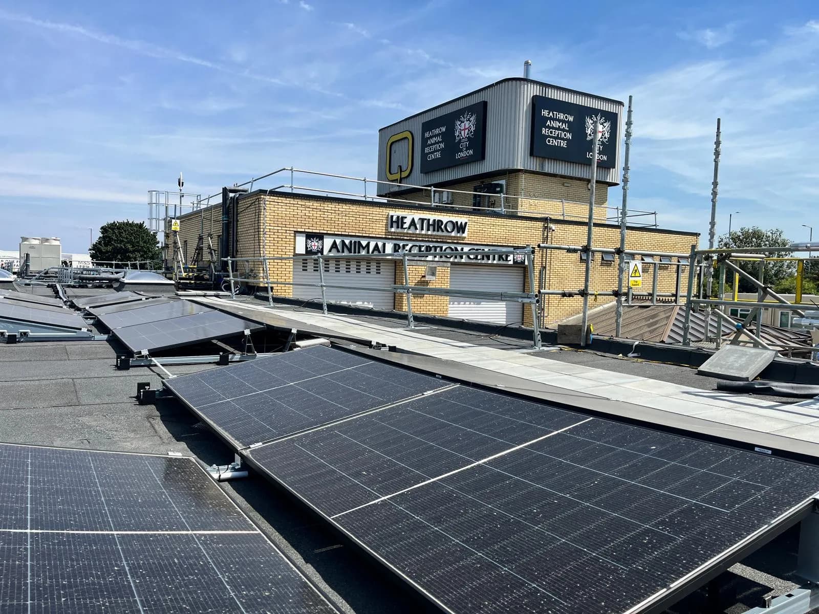 A rooftop view of a brick building with signs for the Heathrow Animal Reception Centre, surrounded by solar panels and scaffolding under a clear sky.