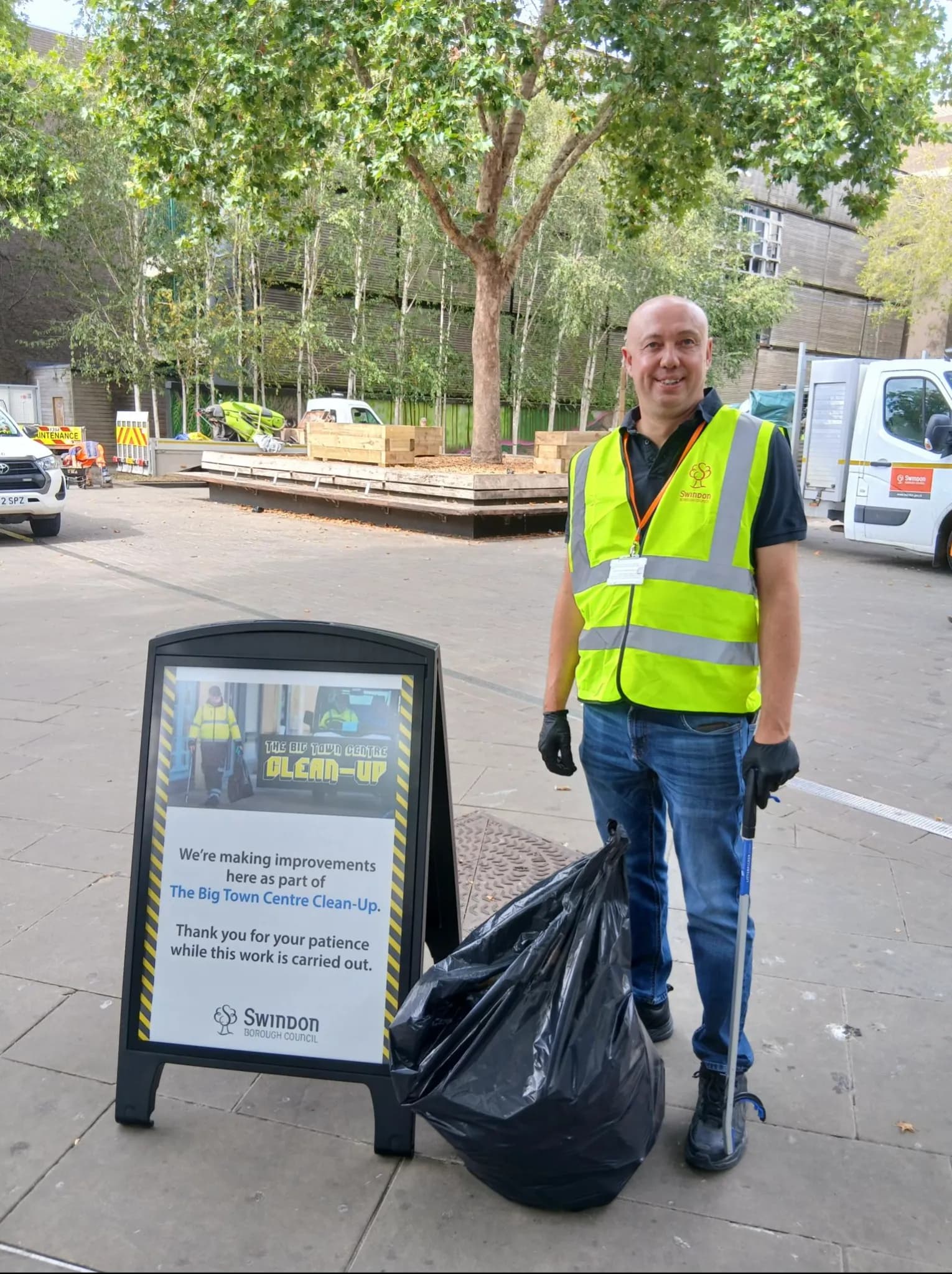 A man in a neon yellow vest and jeans stands next to a black trash bag and a sign for "The Big Town Centre Clean-Up."