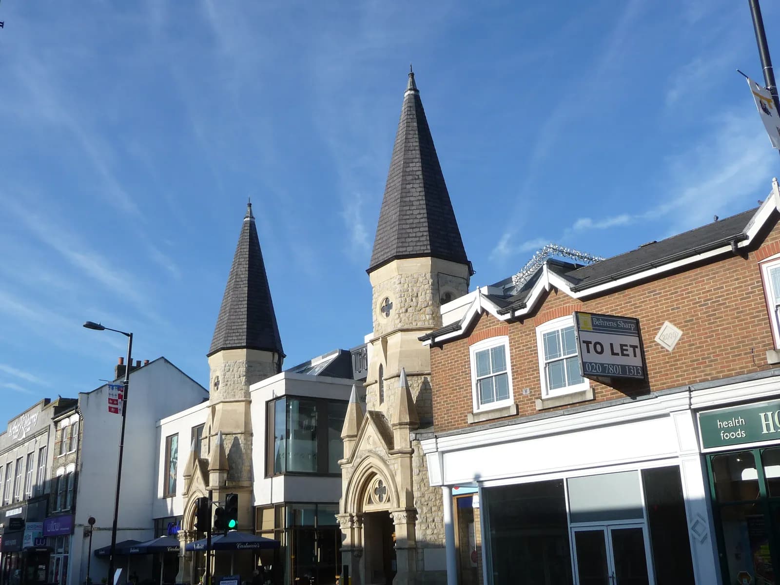 A street scene with various buildings, including one with two prominent spires, under a blue sky with wispy clouds.
