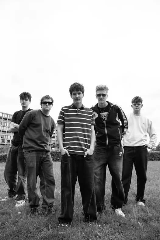 Five male teenagers are standing outdoors in black and white, one in a striped shirt in the middle.