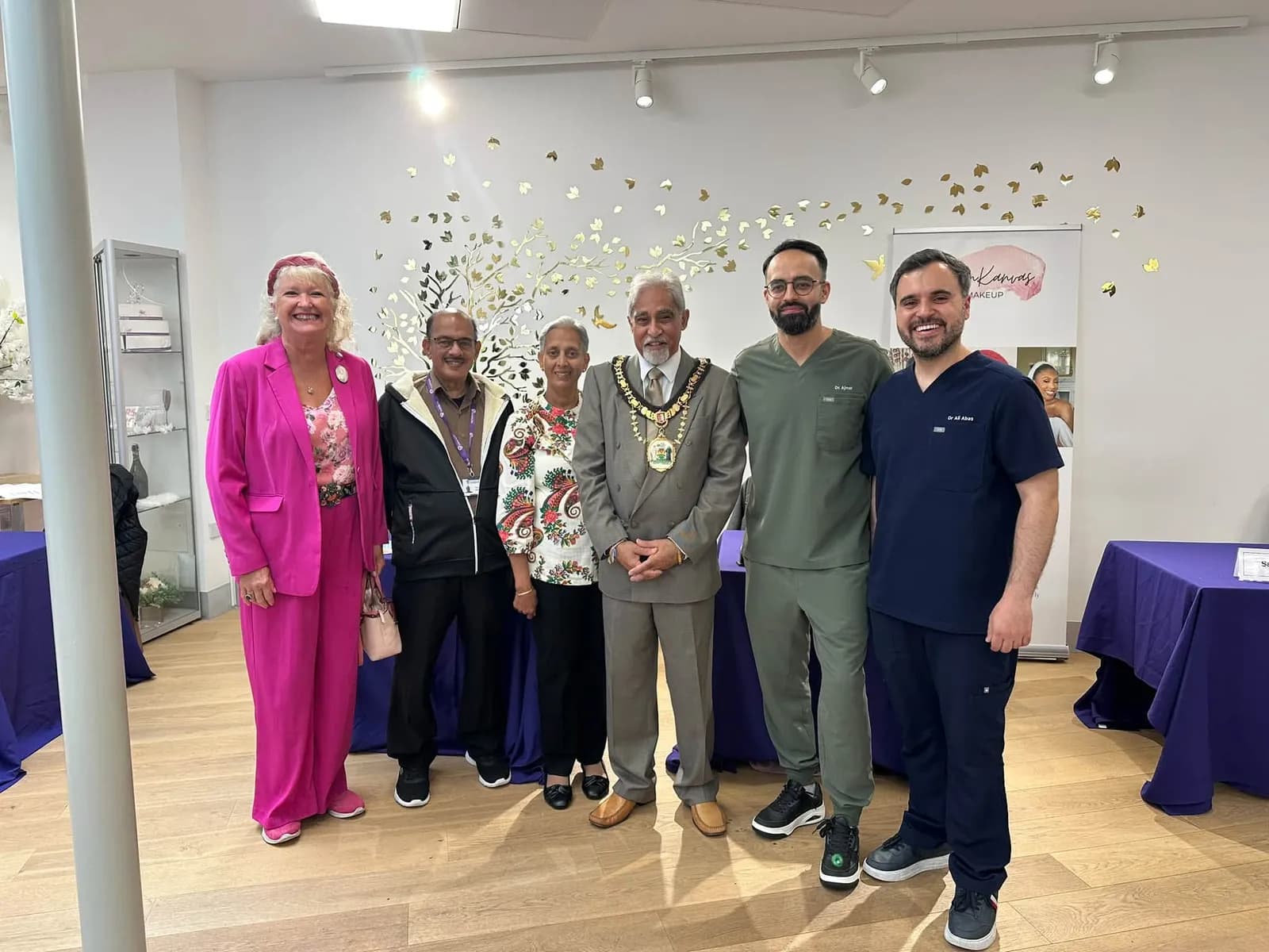 Seven people pose for a group photo in an event space, with three men on the right wearing medical scrubs and a man in the center wearing a ceremonial chain over a suit.