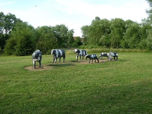 A herd of black and white cow statues stands in a grassy field surrounded by trees under a clear sky.