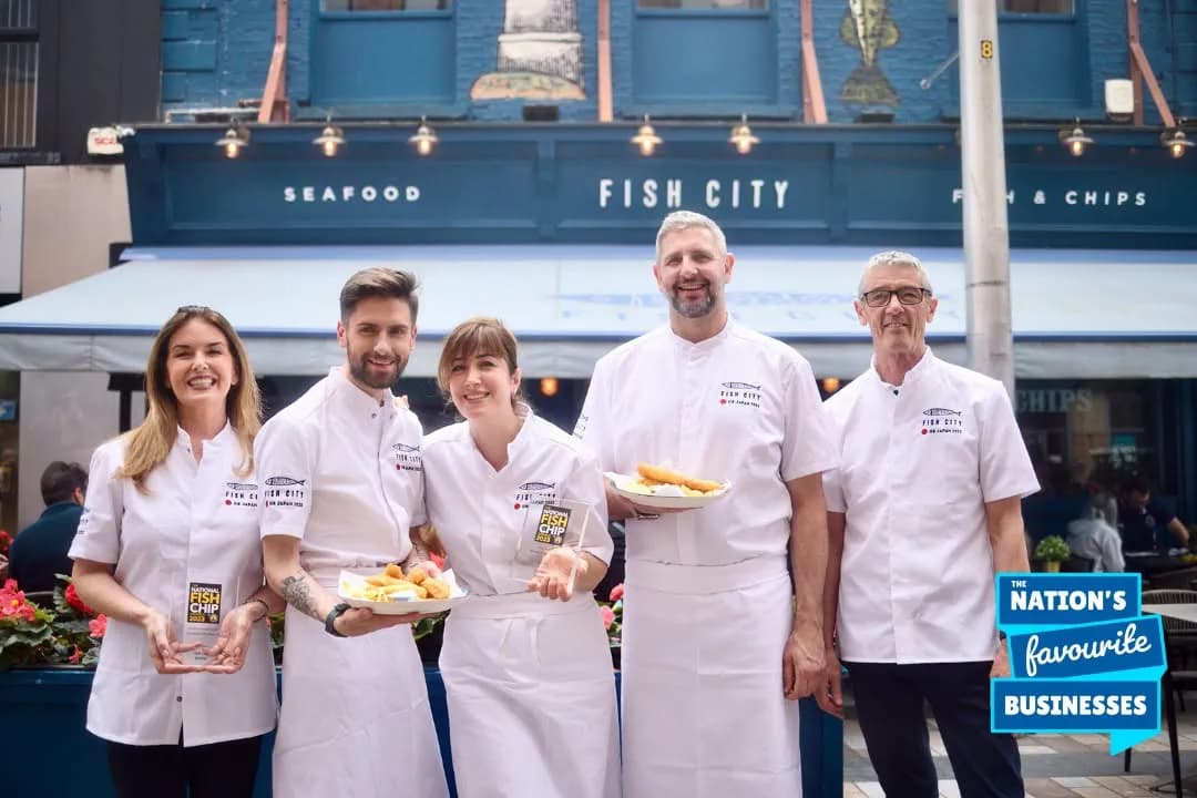 Five chefs from Fish City, a fish and chips restaurant, stand outside their establishment, holding plates of food and an award trophy.