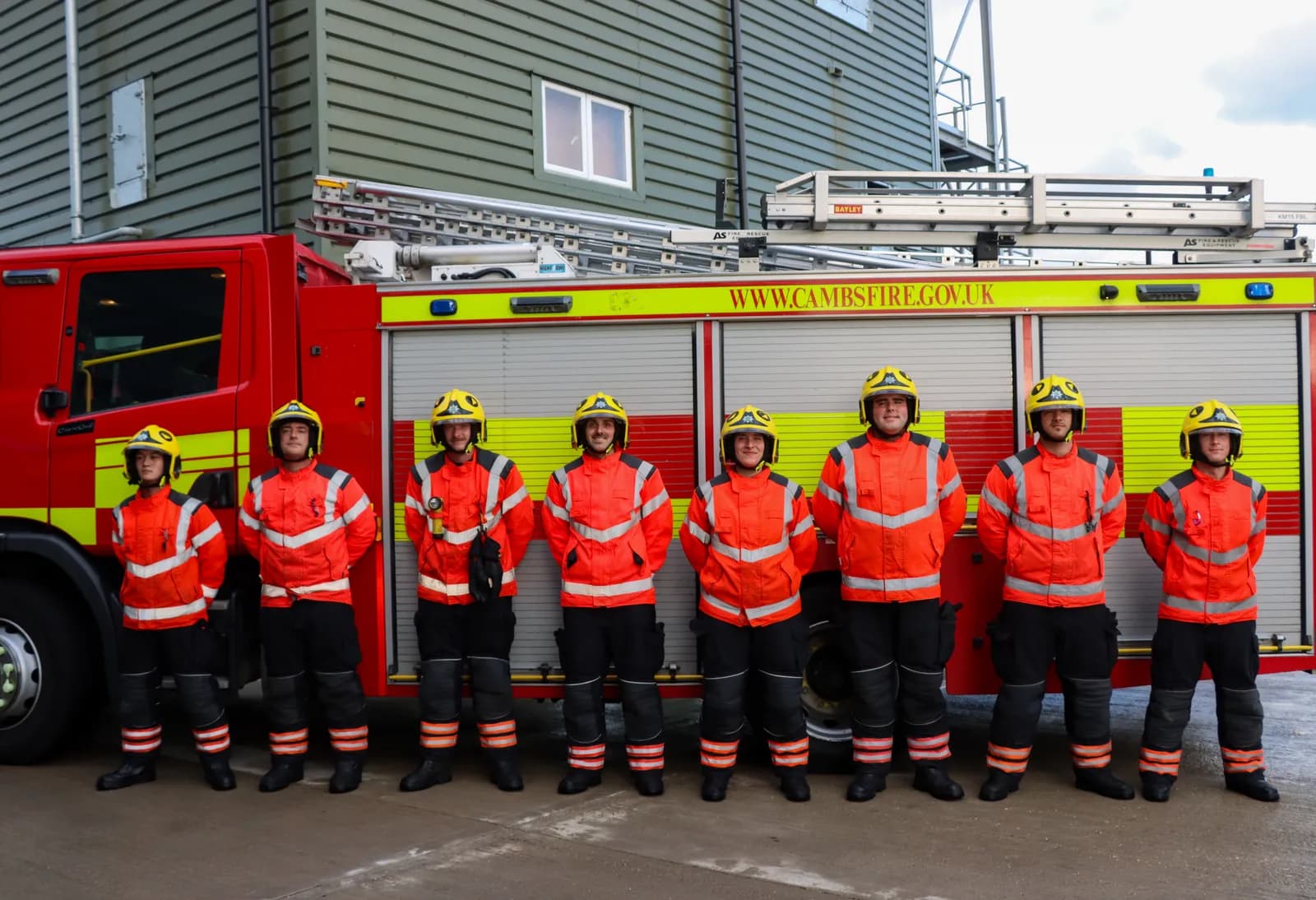 A line of eight firefighters in full orange and black uniforms with yellow helmets stand in front of a red fire truck and a light green building.