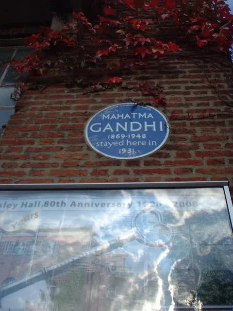 A blue plaque honoring Mahatma Gandhi is affixed to a brick wall with red ivy, and below it is a blurry reflective sign.