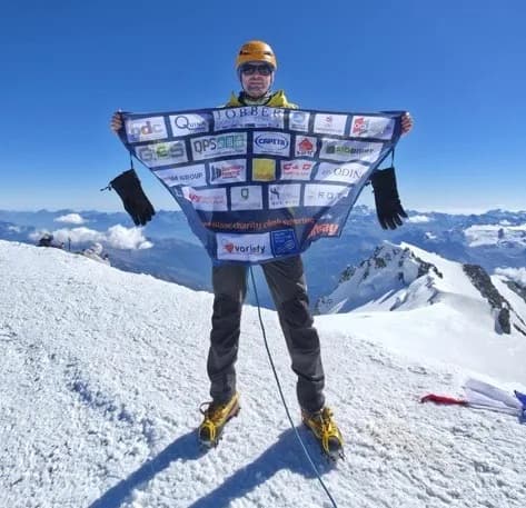 A person wearing a helmet and sunglasses stands on a snow-covered mountain peak, holding up a banner with various logos.