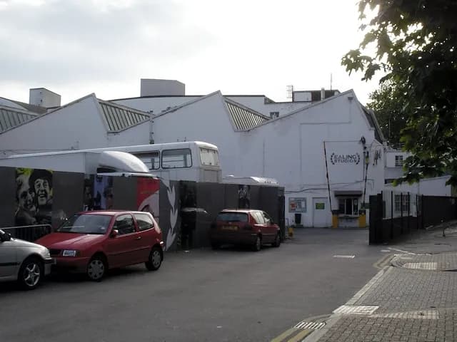 A wide shot of Ealing Studios on a cloudy day, showing its distinctive sawtooth roof, a parked white bus, and several cars, with foliage partially obscuring the top right.