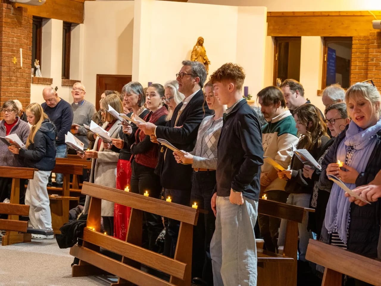People of various ages standing in a church, holding songbooks and some holding lit candles, with wooden pews in the foreground.