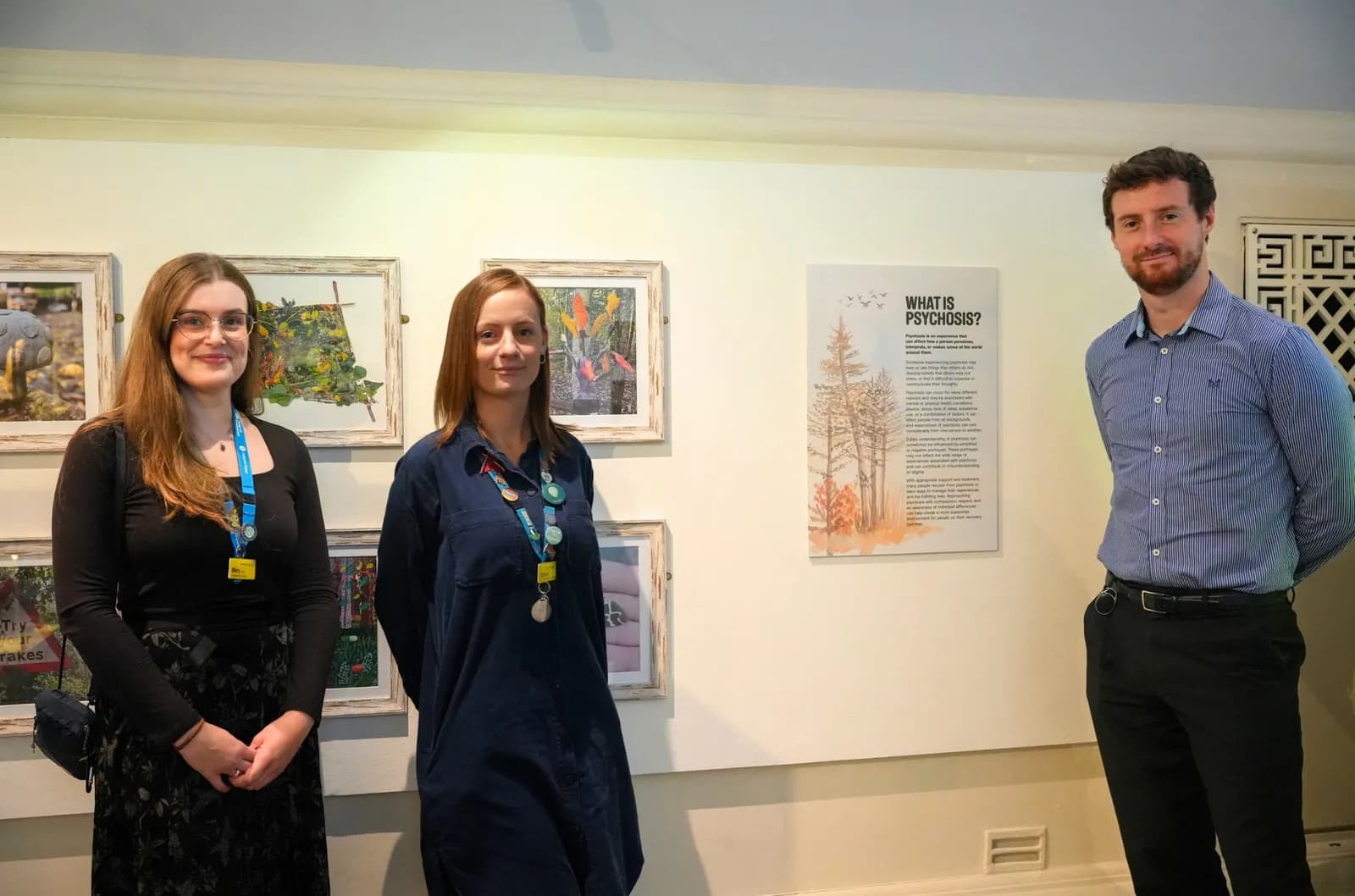 Three people, two women and one man, stand in a room with framed artwork and a sign asking "WHAT IS PSYCHOSIS?" on the wall behind them.