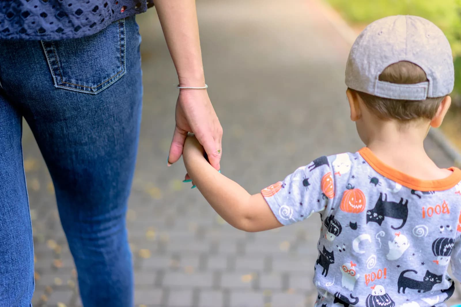 A woman in blue jeans and a blue top holds the hand of a small child in a grey hat and a Halloween-themed shirt with pumpkins, ghosts, and black cats.