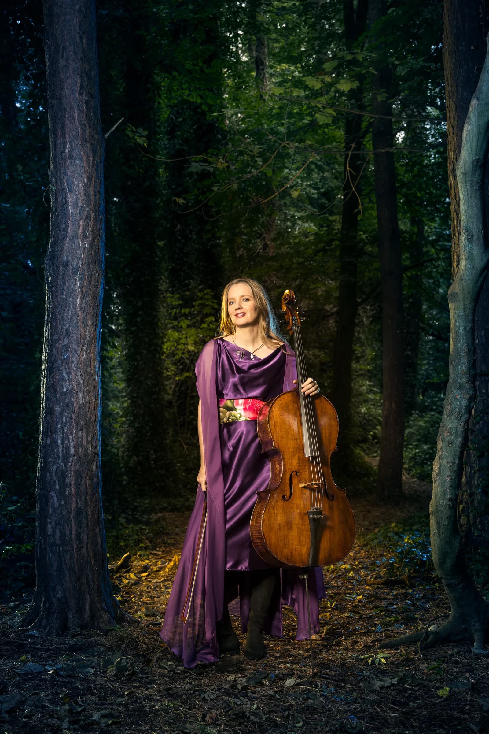 A woman in a flowing purple gown holding a cello while standing in a dimly lit forest.