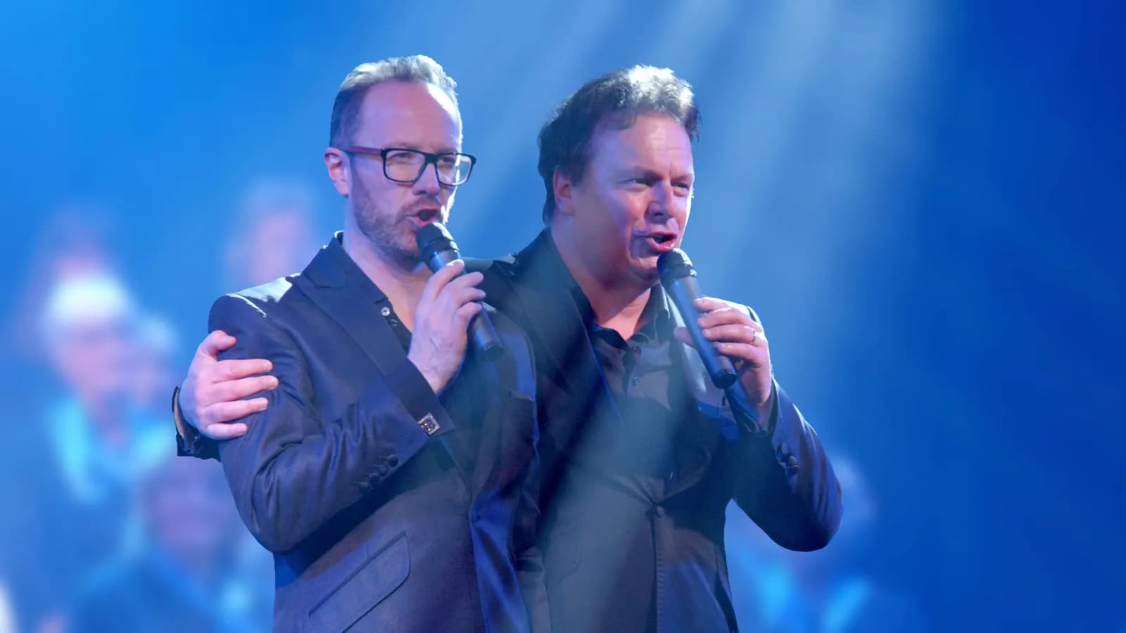 Two men, one with glasses and a beard, the other with dark hair, are singing into microphones on a stage under blue lighting, with the man with glasses holding the other man by the shoulder.