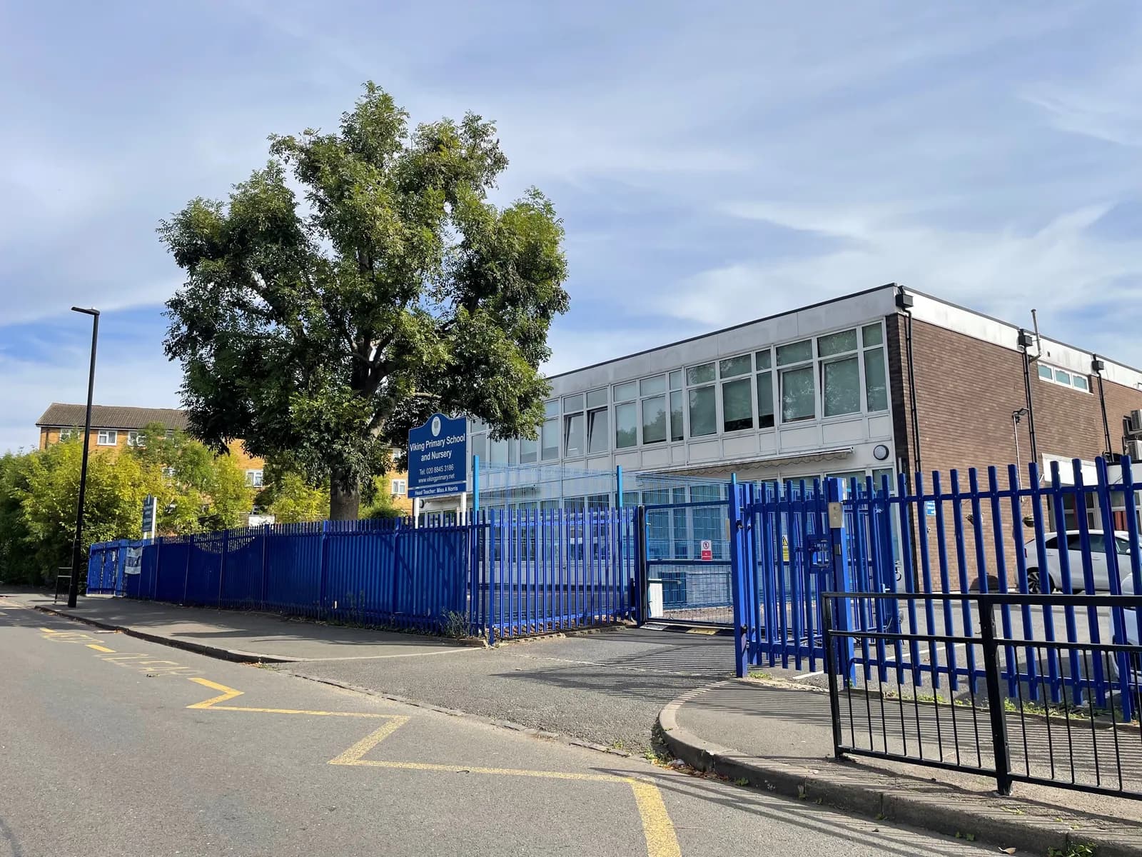 A large building with many windows and a brick facade, behind a tall blue fence, with a prominent tree to the left and a street in the foreground.