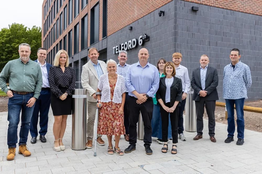 A group of diverse people stand smiling in front of a modern brick building labeled "TELFORD 6th".