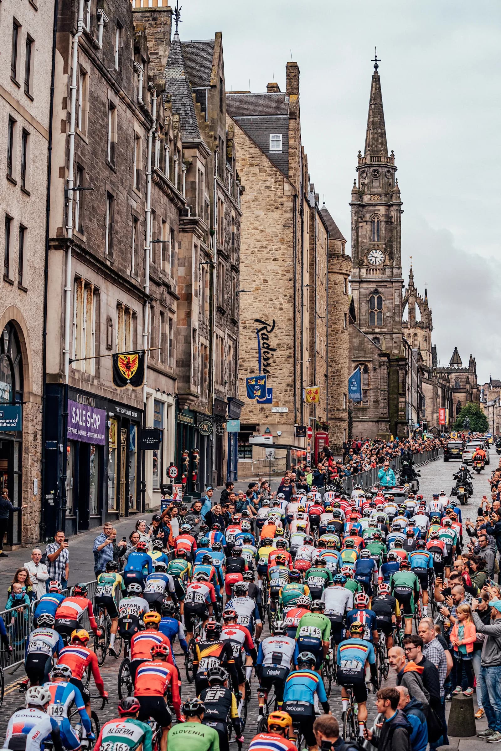 A large group of cyclists in colorful jerseys rides down a cobblestone street lined with historic stone buildings and spectators.