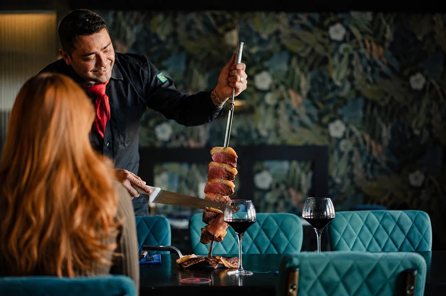 A waiter smiling as he carves a skewer of picanha for a customer at a restaurant table with wine glasses.