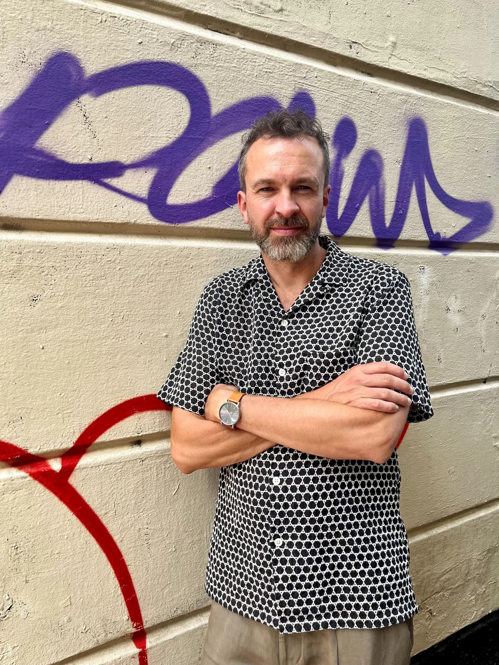 A man with a beard and a patterned shirt stands with his arms crossed against a concrete wall with purple and red graffiti.