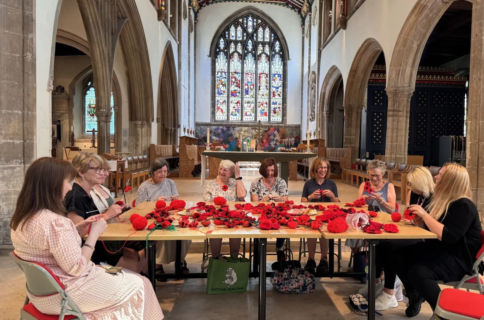 A group of women crafting red poppies at a long table inside a church with high archways and stained glass windows.