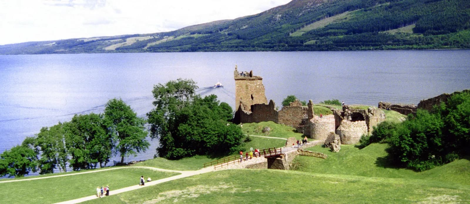 Urquhart Castle ruins on a green peninsula next to a large blue lake with mountains in the background, under a bright sky.