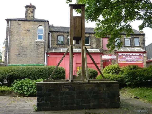 A wooden guillotine stands on a stone base in a paved area, with brick buildings and green bushes in the background under a light sky.