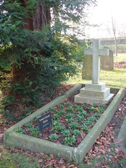 The grave of Lewis Carroll, marked with a small sign and a stone cross at the head, surrounded by greenery and fallen leaves.