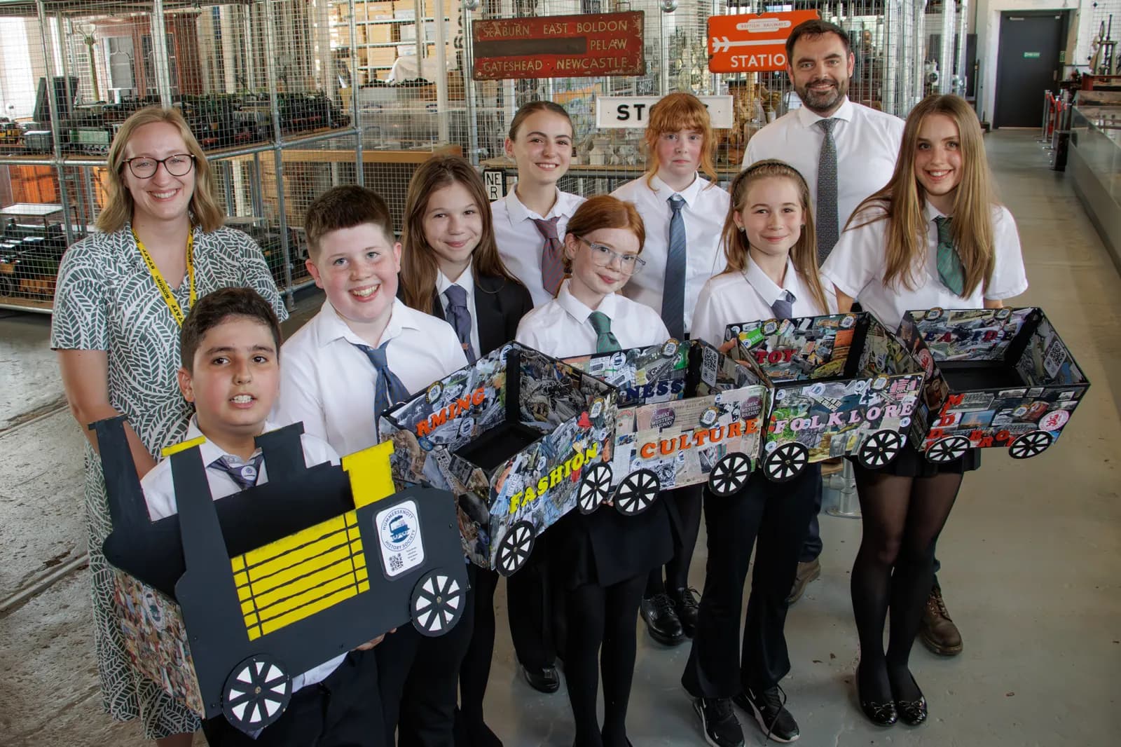 A group of smiling students and two adults standing behind them, holding up a handmade train with various themed wagons.