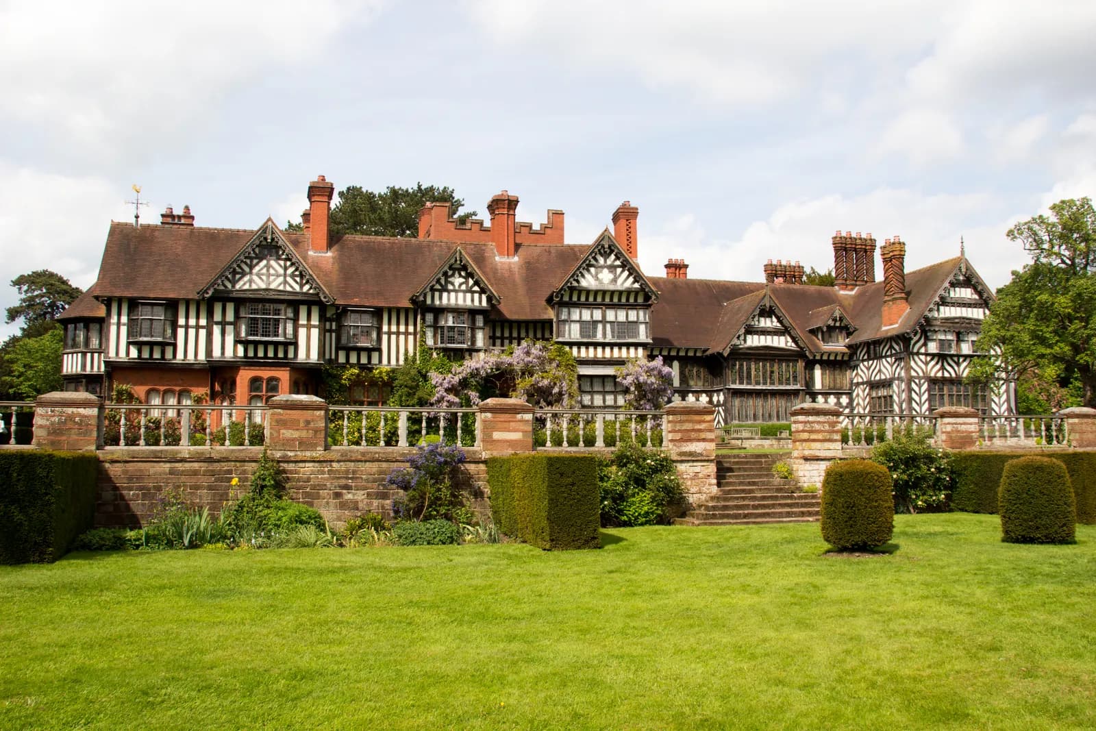 A large, historic house with dark timber framing over white walls and a reddish-brown roof, surrounded by a manicured green lawn, hedges, and flowering plants.