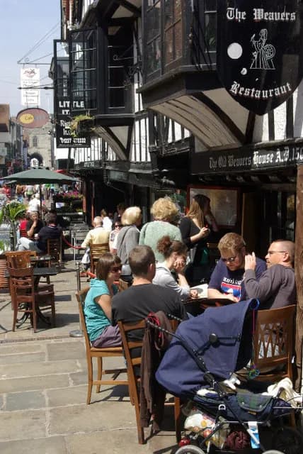 Patrons enjoy an outdoor meal at a restaurant named The Weavers, with half-timbered buildings and a bustling street in the background.