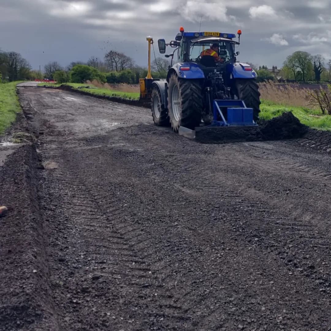 A blue tractor with large tires and an orange-clad driver works on a dirt road, spreading dark material across the foreground.