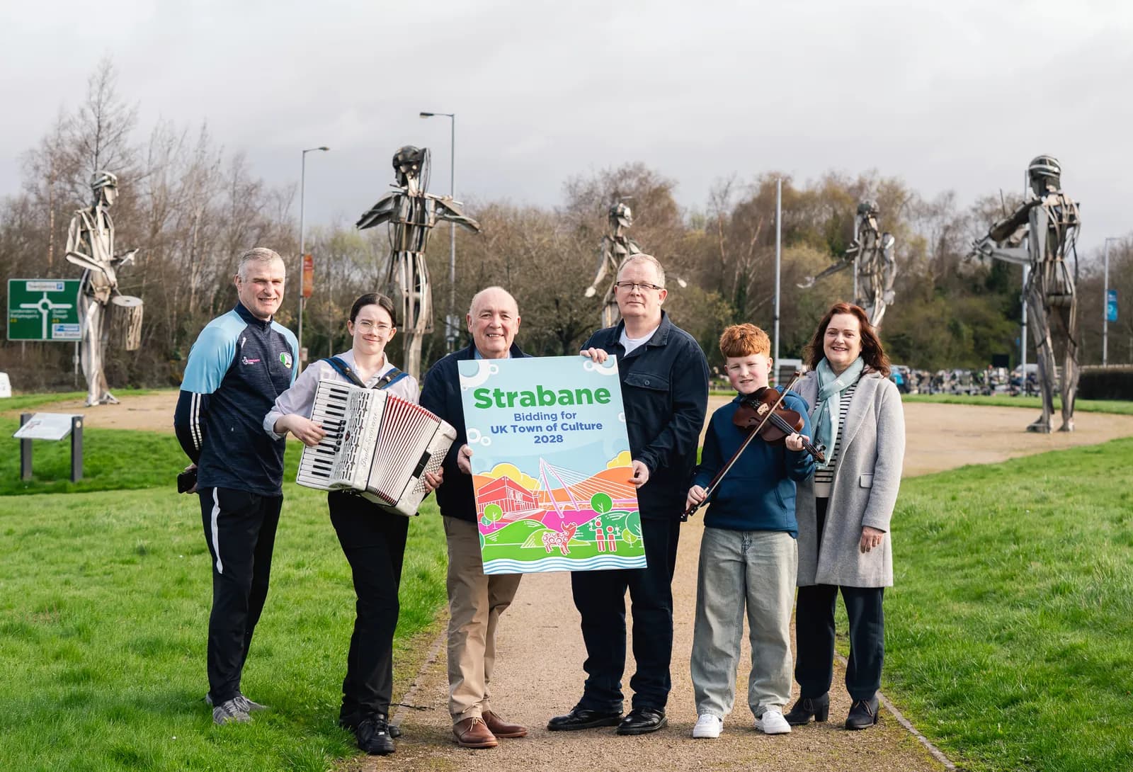 A group of six people stand on a path outdoors, two holding a large poster that reads "Strabane Bidding for UK Town of Culture 2028," a young woman holding an accordion, and a young boy holding a violin and bow.