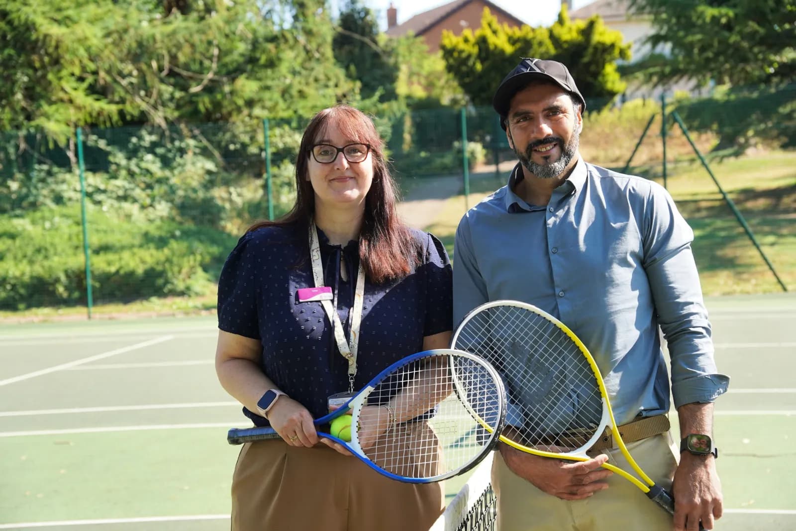 A woman and a man standing side-by-side on a tennis court, both holding tennis rackets and smiling at the camera.