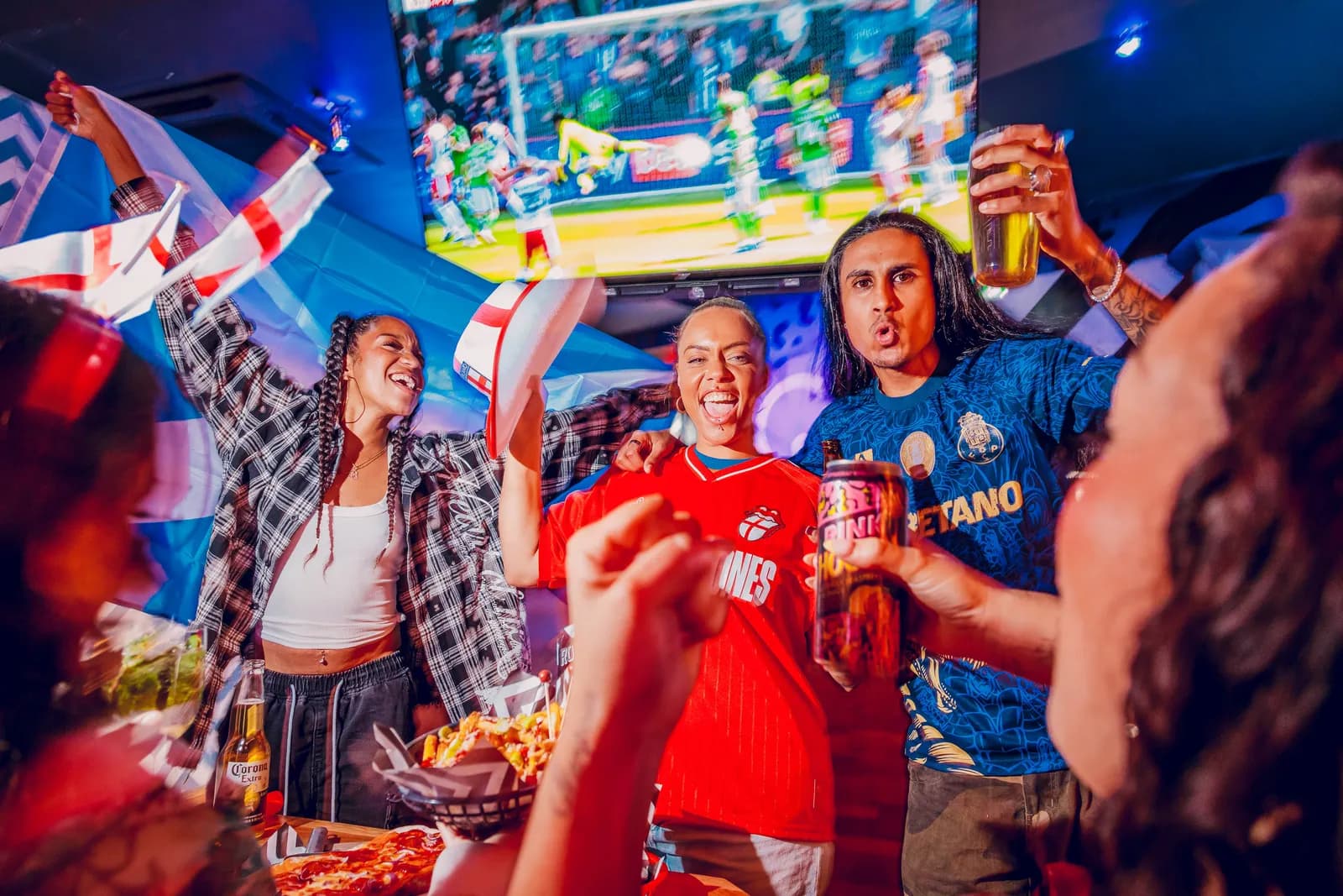 A group of cheerful people holding drinks and flags, celebrating while watching a sports match on a large screen in a dimly lit setting.