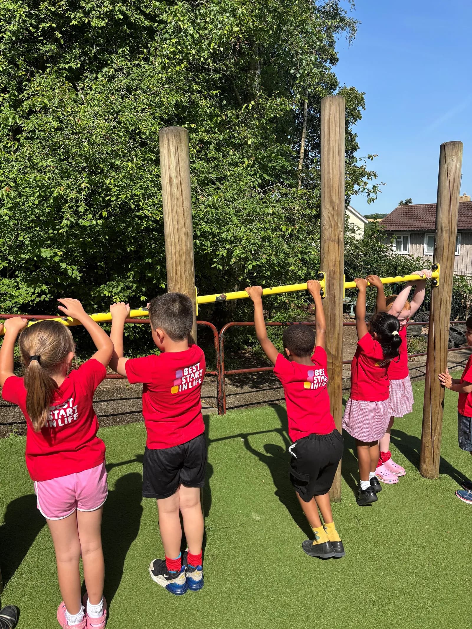 Children in red t-shirts and shorts or skirts hold onto a yellow bar in an outdoor play area.